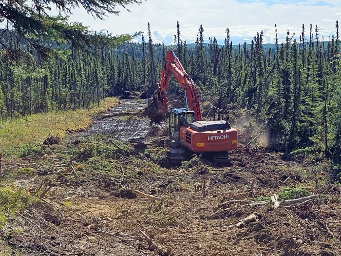 An excavator spreads vegetative material on a dozer fire break in a forested area.