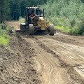 A motor grader smooths a dirt road in a forested area.