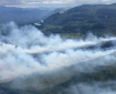 Smoke billows up from two fingers of a fire running parallel to each other. A hill and a river are in the background.