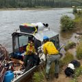 Two people waring yellow shirts and green pants load wildland firefighting equipment onto an airboat on the banks of a river. A man stands on the river bank while handing a woman on the boat white hose.