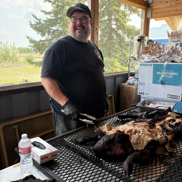 A large smiling man stands on a porch in front of a grill cooking a charred pig with its head still attached.