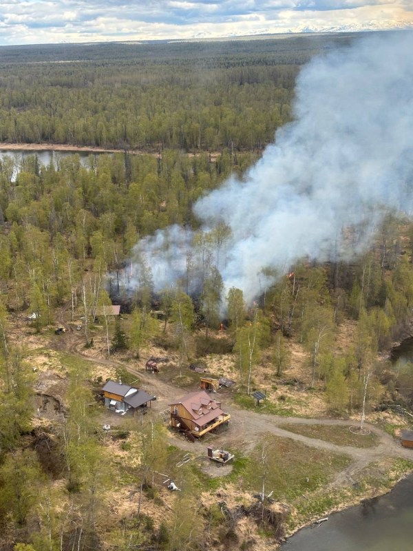 Photo of Yentna Fire burning in grass, hardwoods, and black spruce. Structures are nearby.