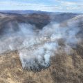 Smoke drifts up from a blackened, burned area along a ridgeline.
