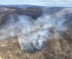 Smoke drifts up from a blackened, burned area along a ridgeline.
