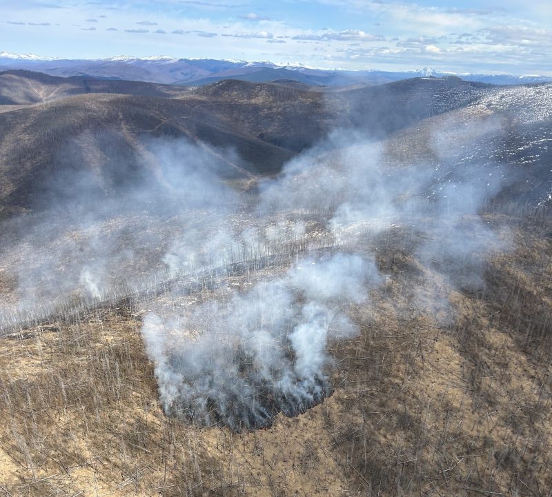 Smoke drifts up from a blackened, burned area along a ridgeline.