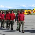 A group of firefighters walk in a single file line across the tarmac at an airport. Two large red and yellow firefighting aircraft are in the background.