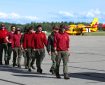 A group of firefighters walk in a single file line across the tarmac at an airport. Two large red and yellow firefighting aircraft are in the background.