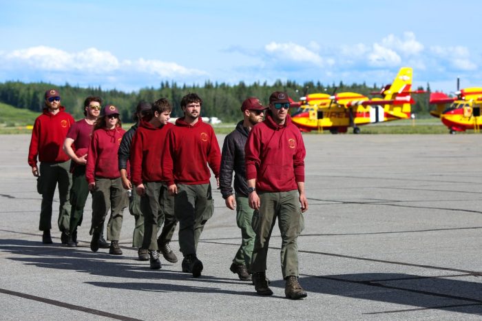 A group of firefighters walk in a single file line across the tarmac at an airport. Two large red and yellow firefighting aircraft are in the background.