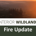 U.S. Wildland Fire Service graphic for a fire update featuring a line of firefighters walking toward a forest fire while the sun sets in the background.