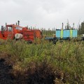 Orange Nodwell with attached trailer carrying a water tank is making its way through dense brush and Black Spruce. The black of the fire is seen in the front left corner of the photo.