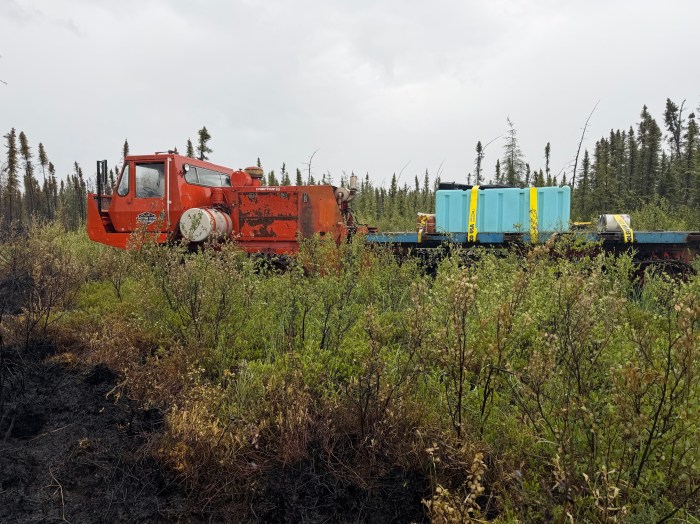 Orange Nodwell with attached trailer carrying a water tank is making its way through dense brush and Black Spruce. The black of the fire is seen in the front left corner of the photo.