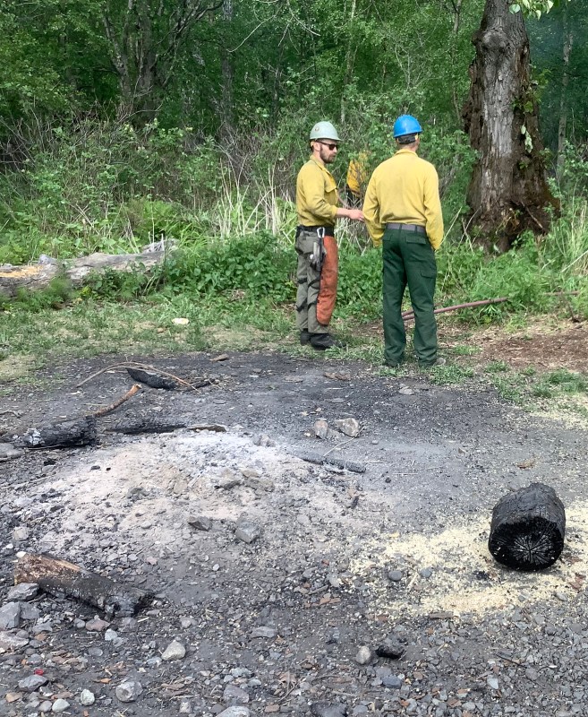 Two wildland firefighters stand next to a campfire and smoldering tree.