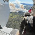 A person wearing a helmet look out from an open airplane door at smoke billowing up from a forest fire.