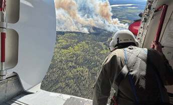 A person wearing a helmet look out from an open airplane door at smoke billowing up from a forest fire.