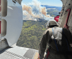 A person wearing a helmet look out from an open airplane door at smoke billowing up from a forest fire.