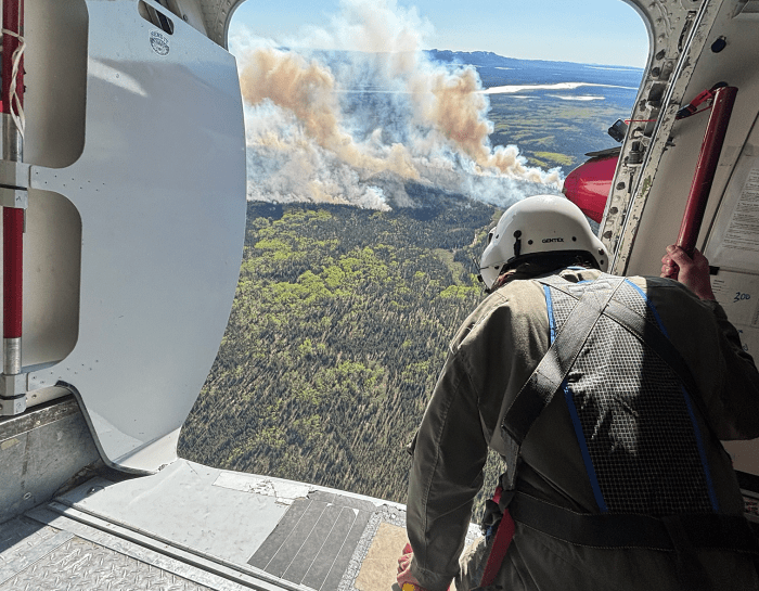 A person wearing a helmet look out from an open airplane door at smoke billowing up from a forest fire.