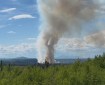 A smoke column rises above a forested area on a sunny, blue-sky day with mountains in the background.