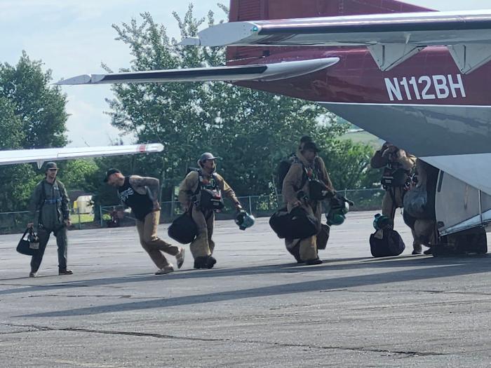 Smokejumpers dressed in fire jumpsuits and helmets carry gear to board CASA aircraft to depart for initial attack.