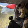 Two people in an airplane look out a doorway at smoke drifting up from a wildfire.