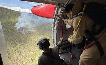 Two people in an airplane look out a doorway at smoke drifting up from a wildfire.