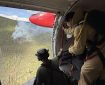 Two people in an airplane look out a doorway at smoke drifting up from a wildfire.