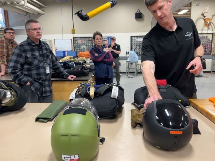 A man touches a black helmet while talking to another man. Two women and two other men listen intently in the background.