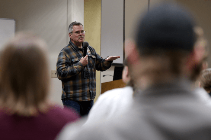 A smiling man gestures while talking into a microphone in front of a group of people.