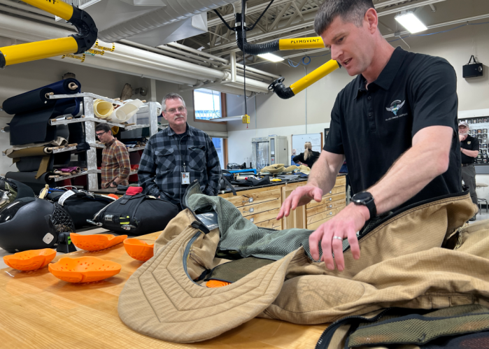A man gestures to a yellow padded suit while another man listens to him talk.