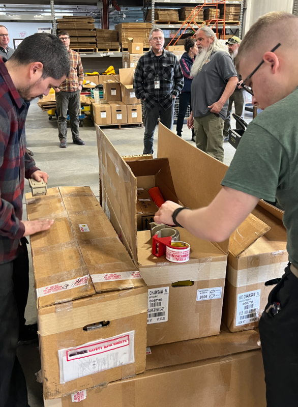 Two men tape up card board boxes while other people gather in the background.