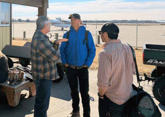 Three men talking with airplanes sitting on a ramp in the background.