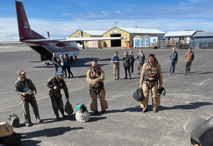 A group of people in flight suits and parachute harnesses stand in a circle while other people stand in the background talking to each other. A red and white plane sits in the background.