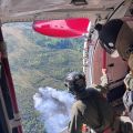 Two people look out from an open airplane door at smoke rising up from a forest fire below.