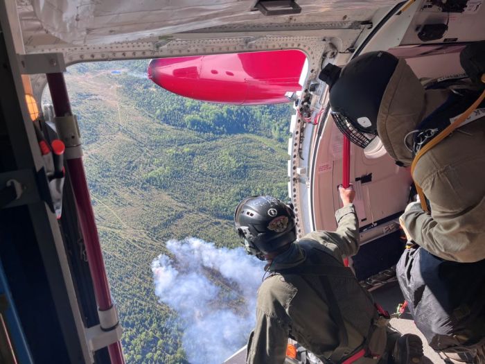 Two people look out from an open airplane door at smoke rising up from a forest fire below.