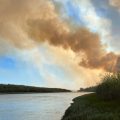 A large column of smoke drifts up from a forest fire burning next to a driver