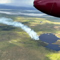 A smoke column drifts up from a forest fire next to a lake. A wide river is in the background.