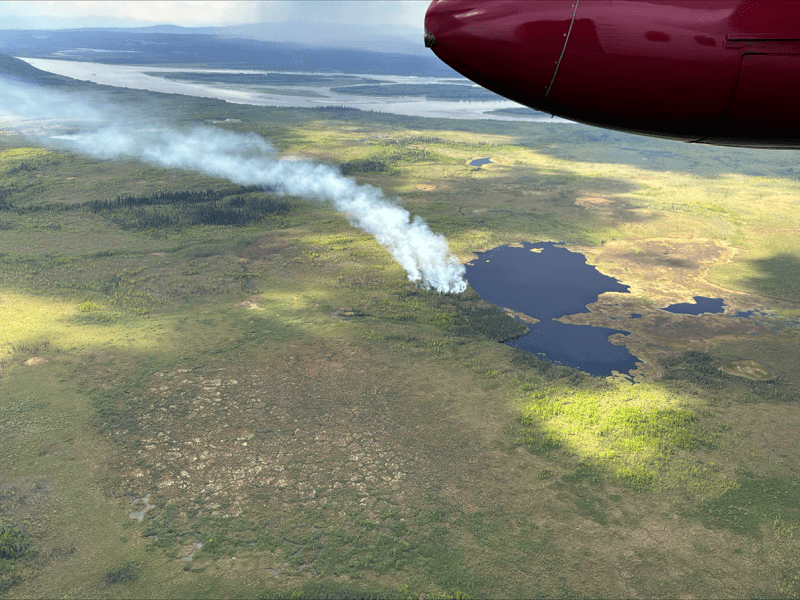 A smoke column drifts up from a forest fire next to a lake. A wide river is in the background.