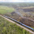 An Alaska Railroad train is on a track stretching from the bottom right corner of the photo to 3/4 of the way toward the upper left. On the left side of the train track, most of the landscape is smoldering from the fire.