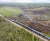 An Alaska Railroad train is on a track stretching from the bottom right corner of the photo to 3/4 of the way toward the upper left. On the left side of the train track, most of the landscape is smoldering from the fire.
