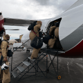 Three people in yellow puffy suits climb steps to get onto a white with yellow and gray stripped airplane.