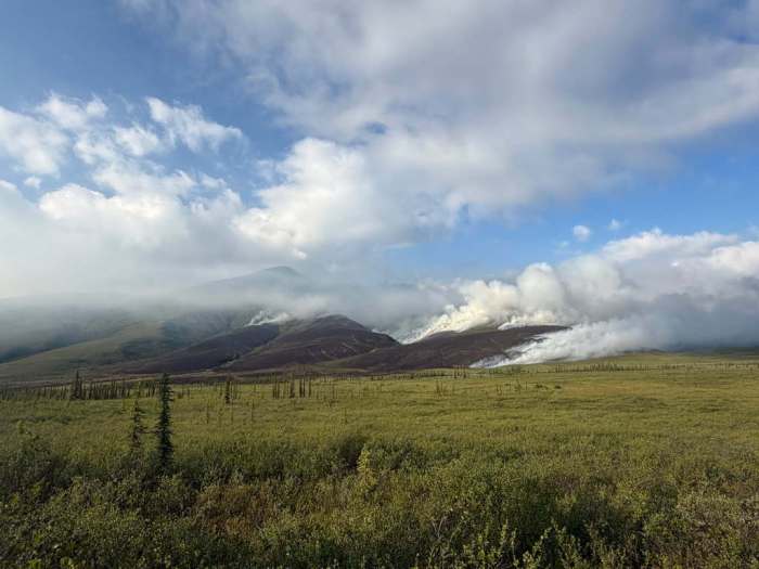 Smoke billows from a low-intensity fire in the mountains with a green tundra foreground.
