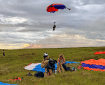 A person decends from the air underneath a blue and orange parachute. Three other people are on the ground including one looking up at the parachuting man and two others either packing up gear or taking off gear.