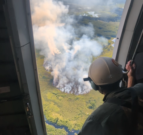 A smokejumper looks from an open airplane door at a smoke rising up from a wildfire below.