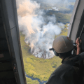 A smokejumper looks from an open airplane door at a smoke rising up from a wildfire below.
