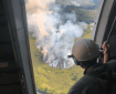 A smokejumper looks from an open airplane door at a smoke rising up from a wildfire below.