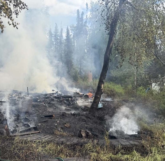 Photo shows smoldering remains of a fire that escaped from a cabin. The ground is black with lots of white smoke and you can see scattered debris smoldering.