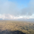 A smokejumper walks along tundra in front of smoke thick enough to obscure views of solar array, fuel tanks and building.