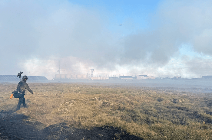A smokejumper walks along tundra in front of smoke thick enough to obscure views of solar array, fuel tanks and building.