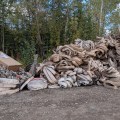A large pile of used fire hose and metal fittings sits on a gravel lot at the edge of a wooded area, awaiting cleaning, inspection, and repacking after wildfire operations.