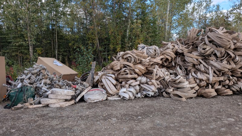 A large pile of used fire hose and metal fittings sits on a gravel lot at the edge of a wooded area, awaiting cleaning, inspection, and repacking after wildfire operations.