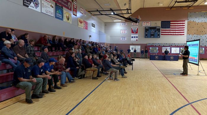 A man stands in front of about 100 people sitting on bleachers at a school gym.